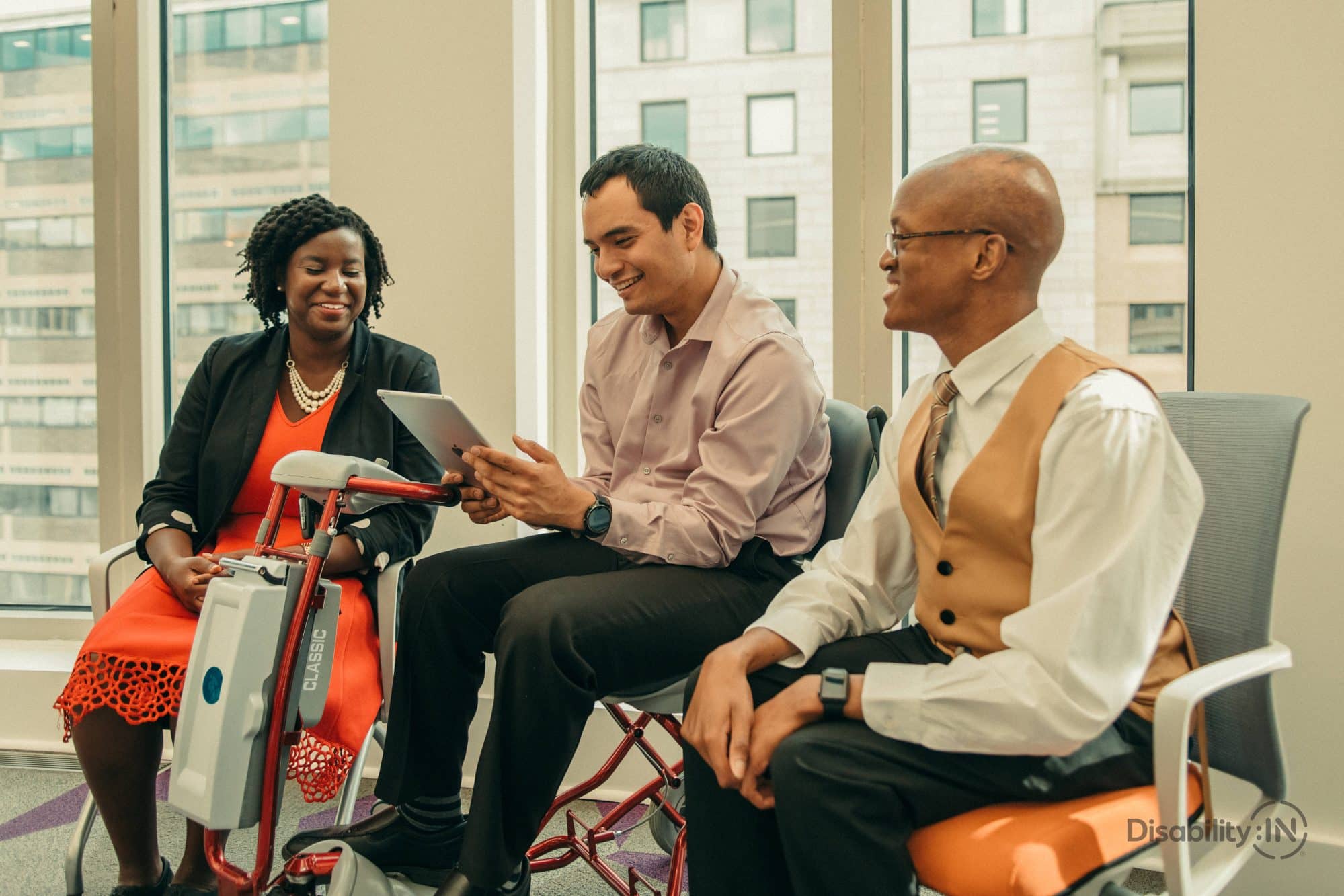 A group of three individuals of diverse backgrounds meeting in a business waiting area; one person uses a mobility scooter while referencing an iPad to a coworker.